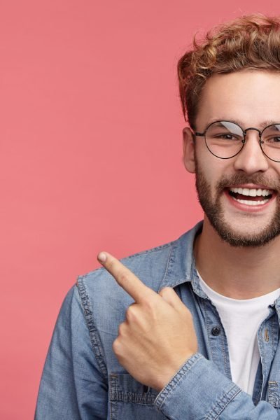 Stylish unshaven man in denim shirt points at copy space on pink wall as shows something pleasant, has smiling look, advertises product. Look over here! People, advertising, emotions concept